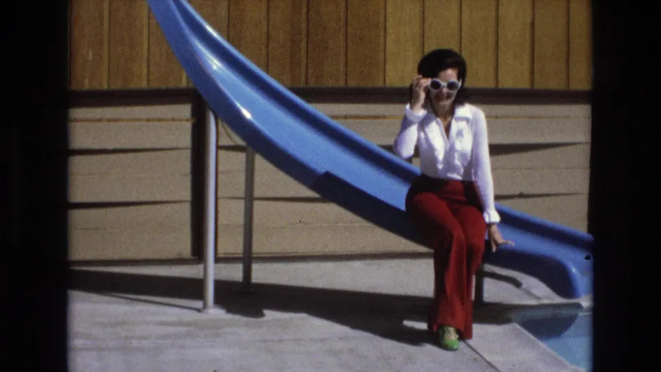 The seated woman remains beside the blue pool slide, holding her white sunglasses while facing the camera in bright daylight.