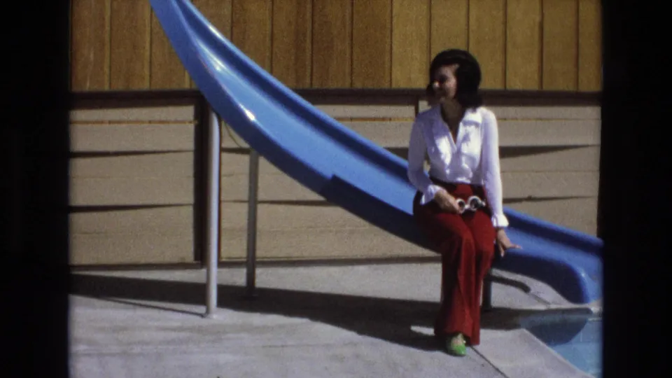 The woman sits at the pool slide base with her white sunglasses lowered in her lap, looking off to the side in the 1973 California home-movie scene.
