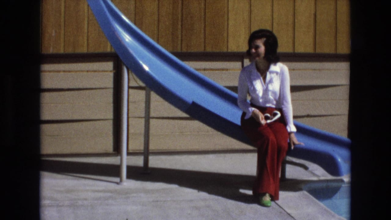 In 1973 Central Coast California home-movie footage, a woman in a white blouse, red trousers, and green shoes sits on a blue playground slide holding sunglasses.