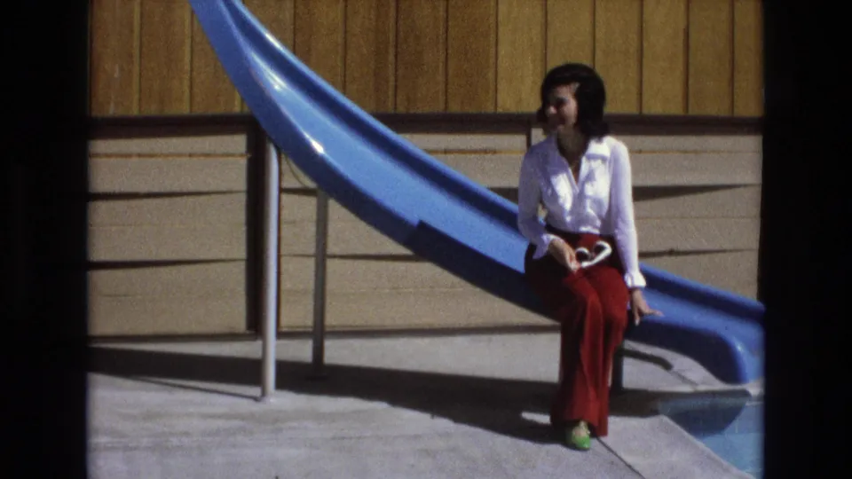 In 1973 Central Coast California home-movie footage, a woman in a white blouse, red trousers, and green shoes sits on a blue playground slide holding sunglasses.