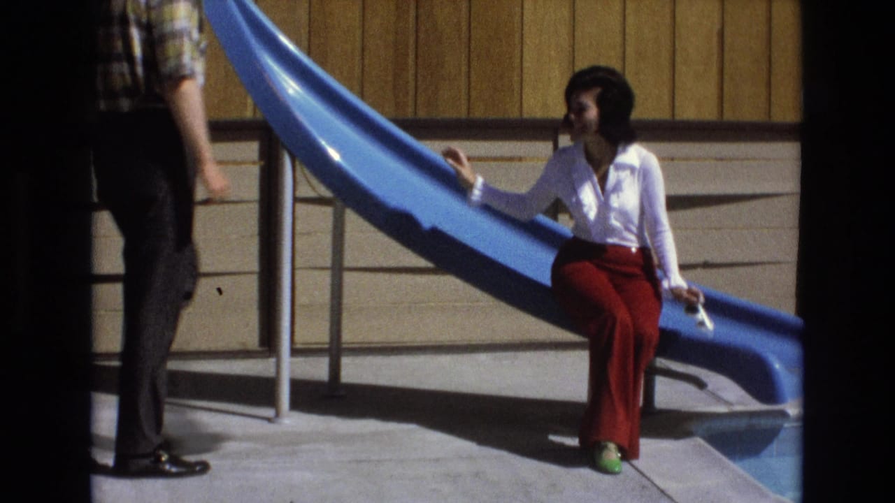 An adult man in a plaid shirt and dark pants approaches the woman seated on the blue slide in the 1973 California home-movie scene.