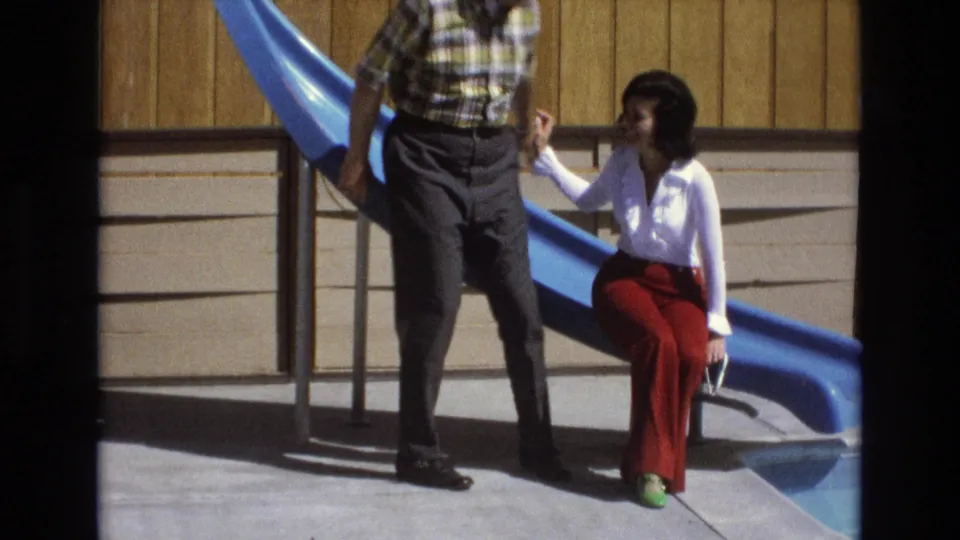The man stands next to the blue slide and reaches toward the seated woman, who looks up from beside the metal railing.
