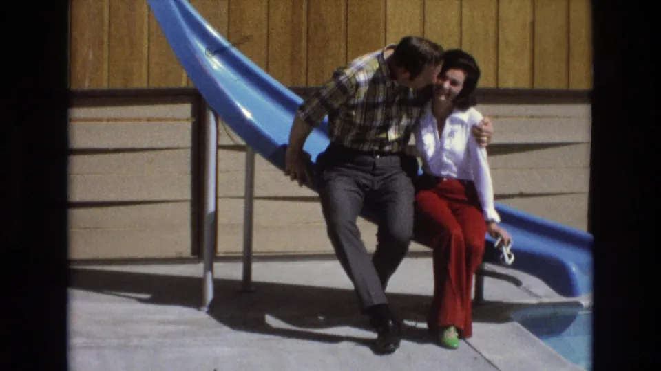 In 1973 Central Coast California home-movie footage, an adult couple sits on the edge of a bright blue slide while the man leans in toward the woman beside a wood-paneled exterior wall.