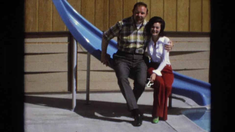 An adult man in a plaid shirt and woman in a white blouse and red trousers smile on a bright blue slide in 1973 Central Coast California home-movie footage.