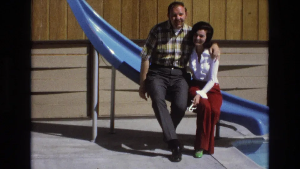 The couple remains seated close together on the blue slide, facing the camera in bright sunlight with a wood-paneled building behind them in the 1973 home-movie frame.