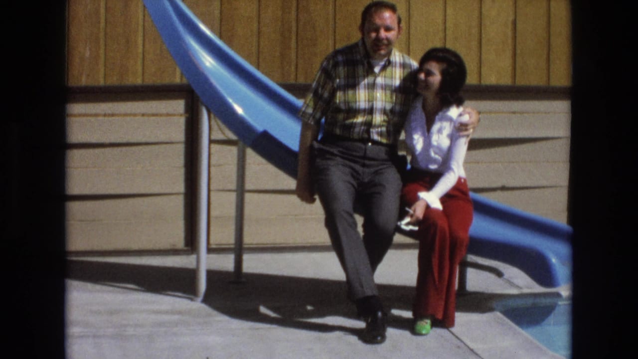 In a 1973 Central Coast California home-movie frame, an adult man in a plaid shirt sits with his arm around a woman in a white blouse and red pants beside a blue slide.