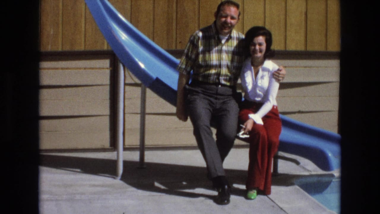 The couple remains seated close together in the 1973 Central Coast California home-movie scene, with the man smiling toward the camera and the woman seated beside the blue slide.