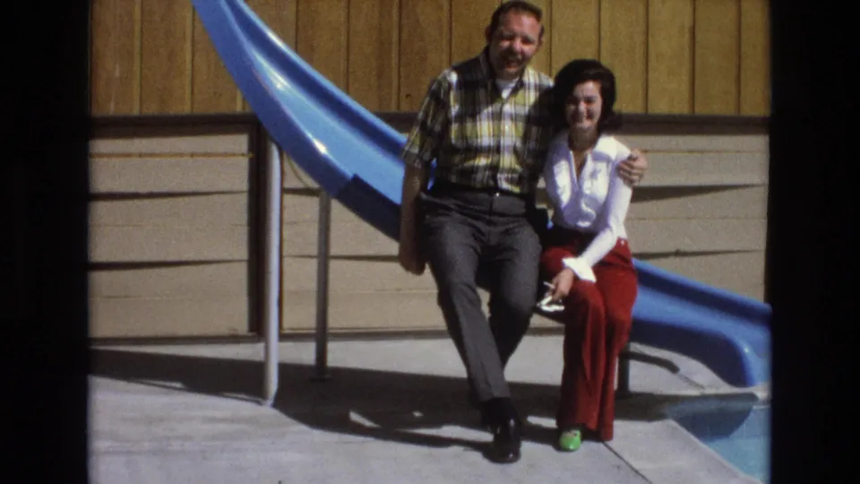 The couple remains seated close together in the 1973 Central Coast California home-movie scene, with the man smiling toward the camera and the woman seated beside the blue slide.