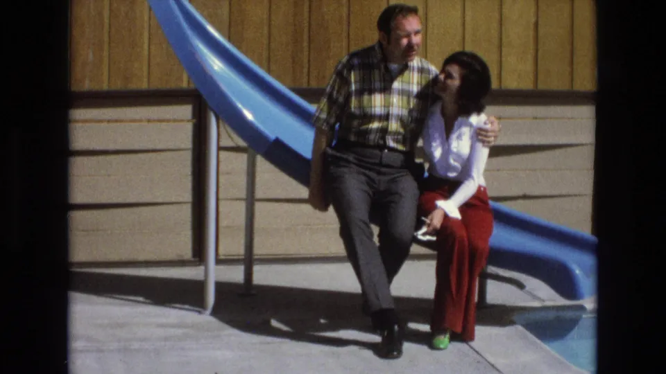 In this 1973 Central Coast California frame, the man and woman sit closely by the blue slide as the woman turns toward him and the sun casts strong shadows on the concrete.