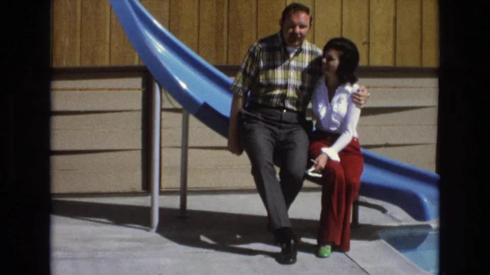 The closing 1973 Central Coast California home-movie frame shows the couple still seated together beside the blue playground slide, framed by the dark edges of the film scan.