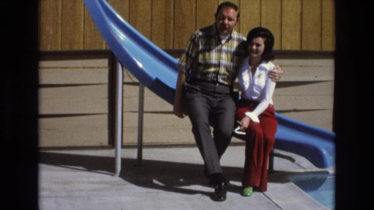 Adult couple sitting close together beside a bright blue slide in sunlight.