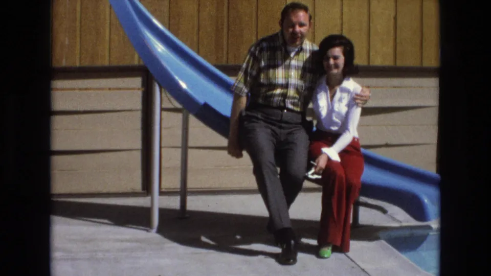 The couple remains seated together beside the blue slide, with the man leaning close and holding the woman as they face the camera in bright outdoor light.