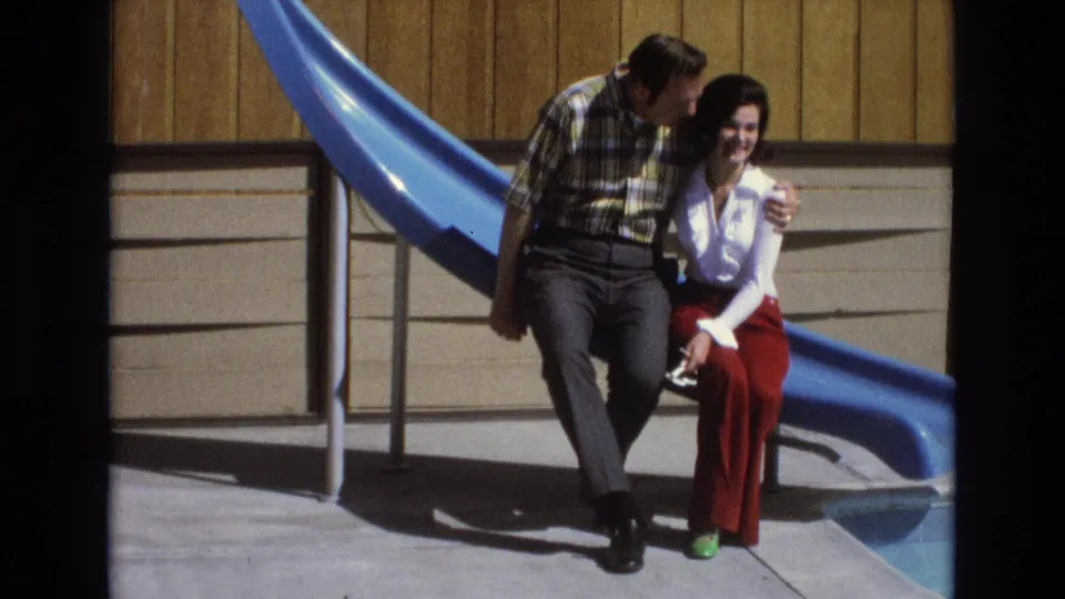 The man leans toward the woman in an affectionate moment while they sit shoulder to shoulder beside the bright blue slide in the 1973 home-movie scene.