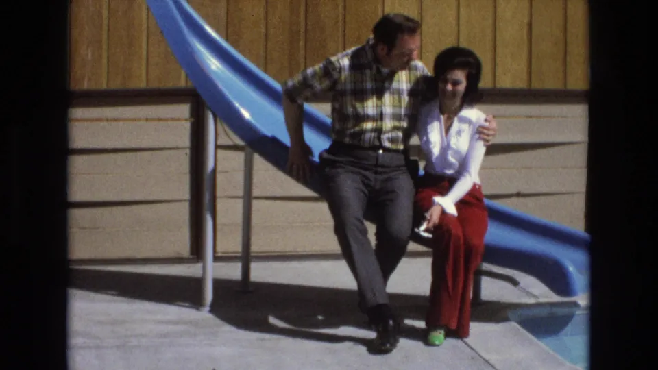 At the end of the clip, the man turns toward the woman while keeping his arm around her as they sit by the blue slide against a wood-paneled wall.