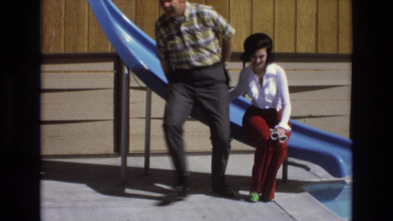 A man in a plaid shirt and dark trousers sits beside a woman in a white blouse and red bell-bottom trousers near a blue pool slide in 1973 Central Coast California home-movie footage.
