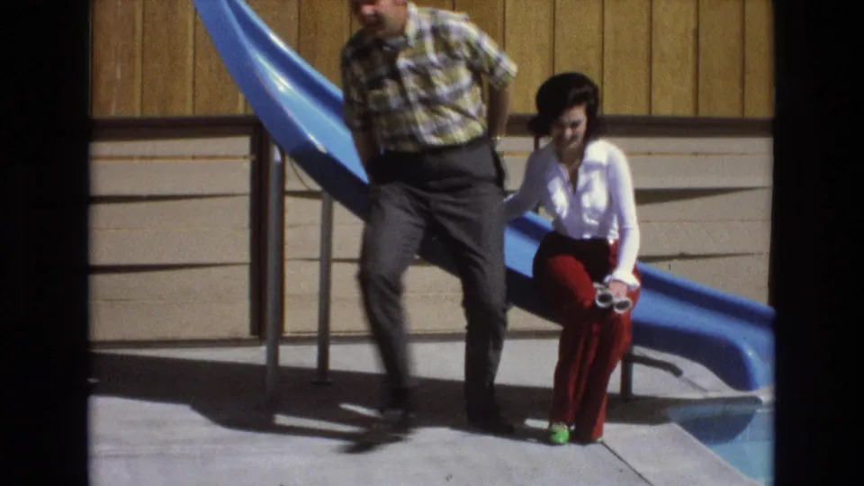 A man in a plaid shirt and dark trousers sits beside a woman in a white blouse and red bell-bottom trousers near a blue pool slide in 1973 Central Coast California home-movie footage.
