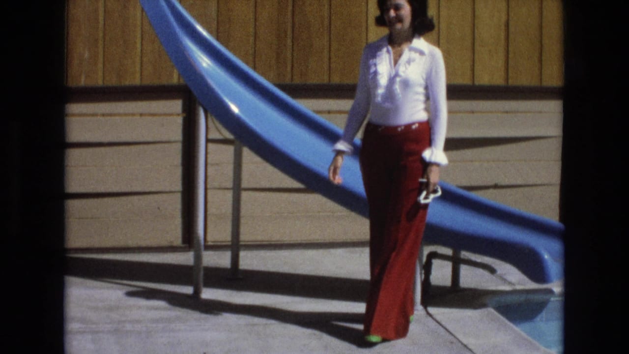 Woman standing beside a blue pool slide while holding shoes.