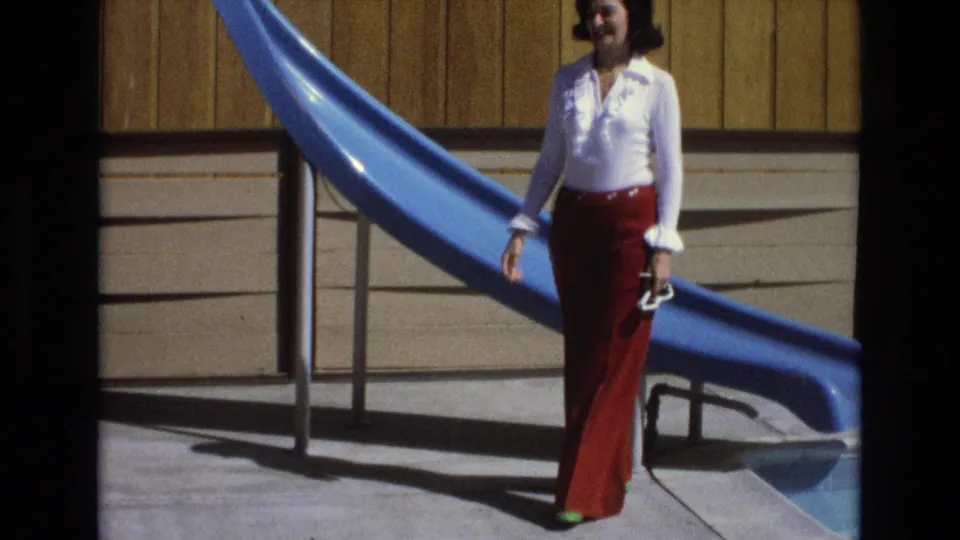 A woman in a white ruffled blouse and red bell-bottom trousers stands beside a blue pool slide, holding shoes in her hand in the sunlit California scene.