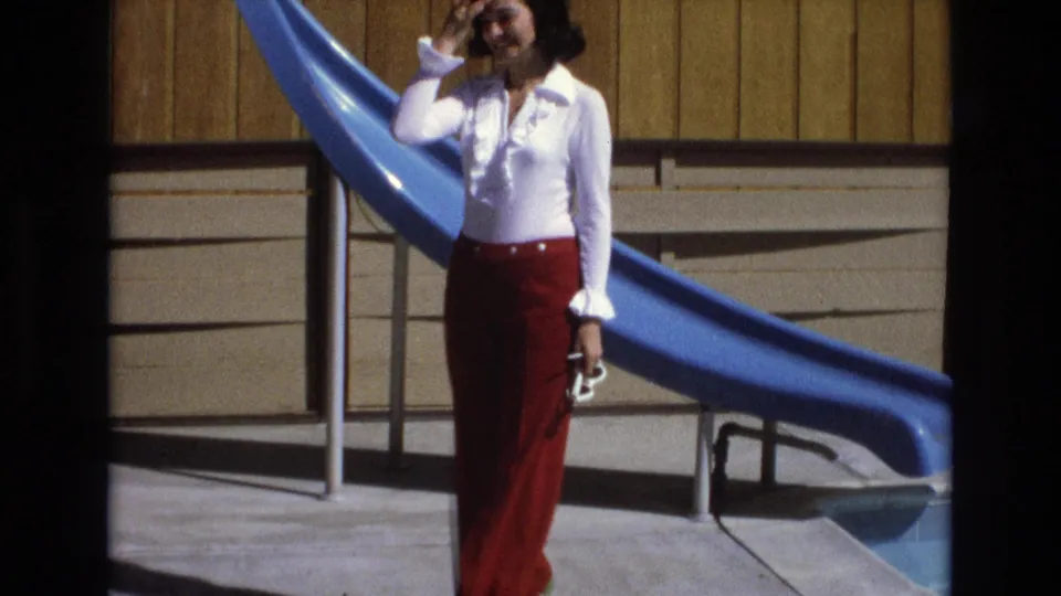The woman stands close to the blue pool slide with one hand raised near her face and shoes in her other hand, wearing 1970s-style red bell-bottom trousers.