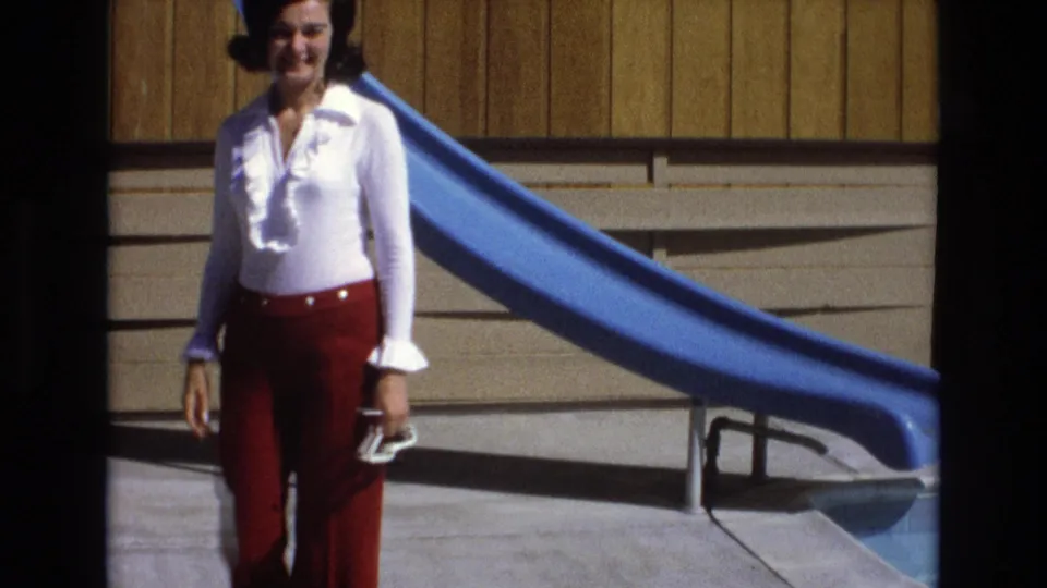 A woman in a white blouse and red bell-bottom trousers walks toward the camera beside the blue pool slide, holding shoes in late-day-looking sunlight.