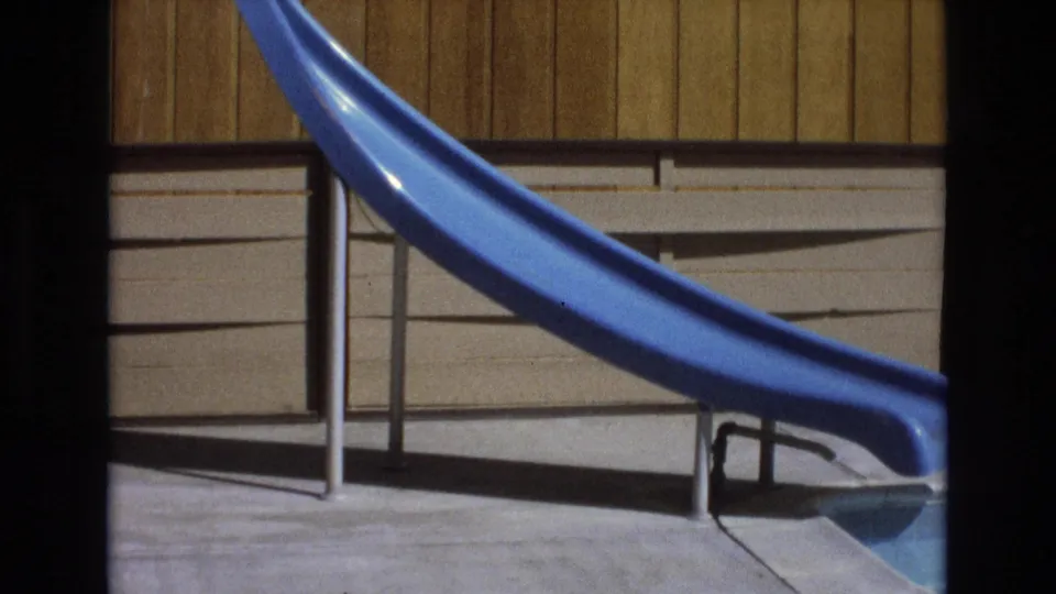 The blue water slide curves across a sunlit poolside area with railings and a building exterior visible behind it.