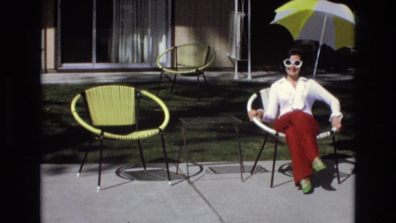 In 1973 Central Coast California home-movie footage, a woman in white sunglasses, a white blouse, red pants, and green shoes sits on a concrete patio near yellow outdoor chairs and a yellow-and-white umbrella.