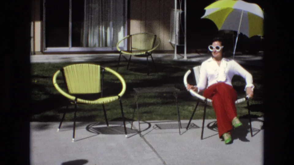 A sunlit 8mm frame shows the woman seated with legs crossed on a concrete patio, framed by yellow patio chairs, a lawn strip, house windows, and the yellow-and-white umbrella.