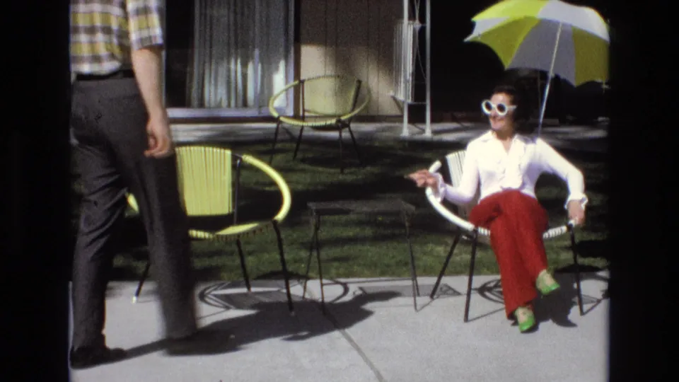 A man in a plaid shirt and dark trousers crosses the foreground while the woman in sunglasses remains seated near yellow patio chairs, lawn, windows, and a striped umbrella.