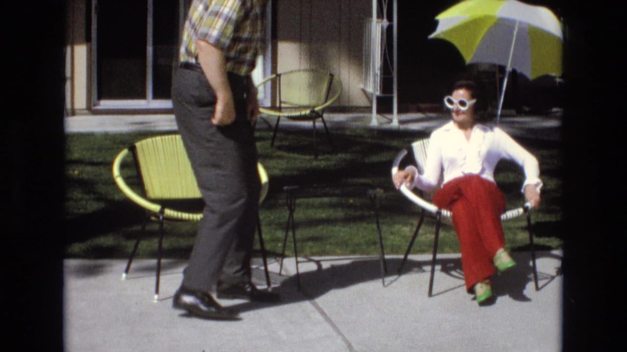 1973 Central Coast California home-movie frame showing a man standing near a seated woman in white sunglasses on a sunny patio with modernist chairs and an umbrella.