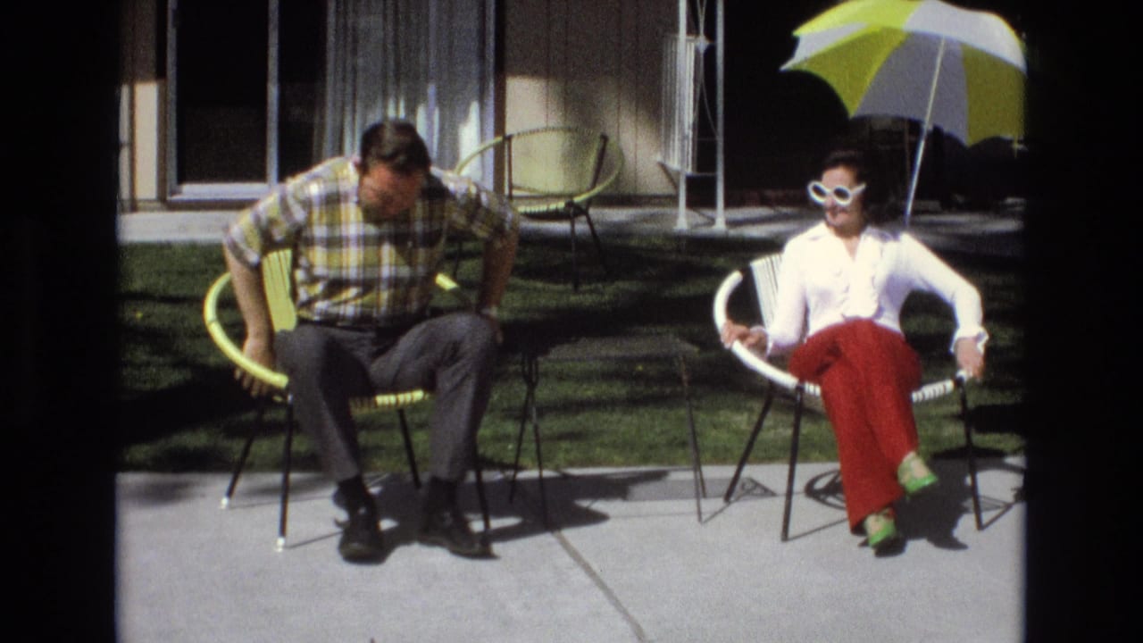 Man lowering himself into a yellow patio chair beside a woman in red pants.