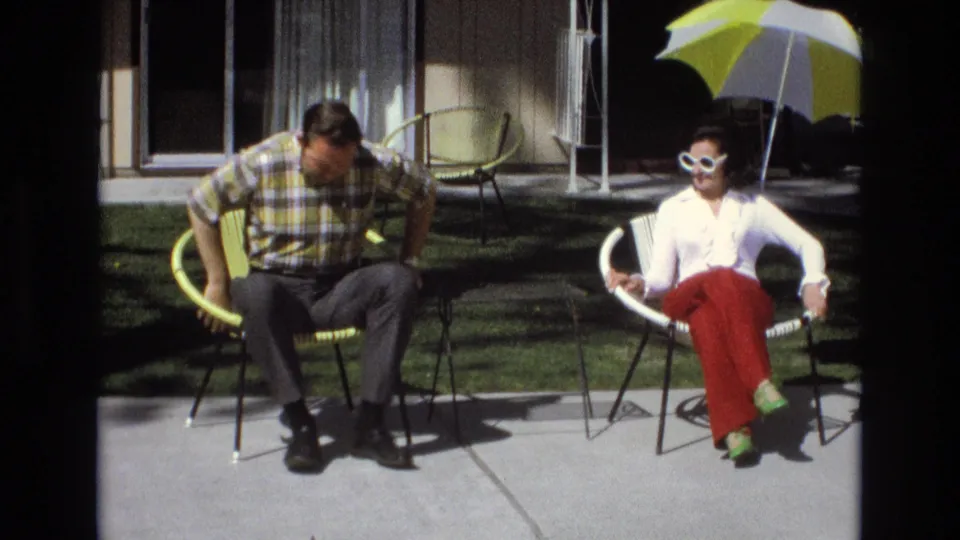 1973 Central Coast California home-movie frame showing a man sitting down in a yellow patio chair beside a woman in red pants and white sunglasses under a yellow-and-white umbrella.