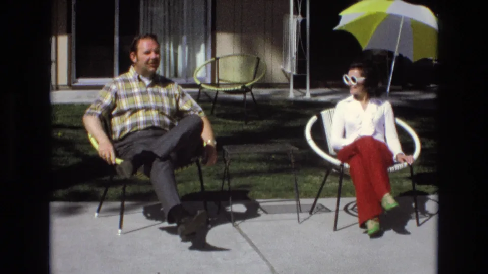 In a 1973 Central Coast California home-movie frame, a man in a plaid shirt and a woman in white sunglasses sit on patio chairs in bright sun.