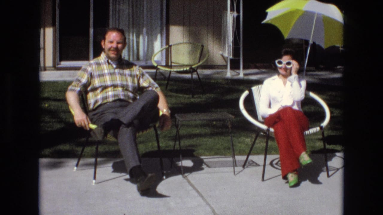 Seated couple on patio with the woman wearing white sunglasses under an umbrella.