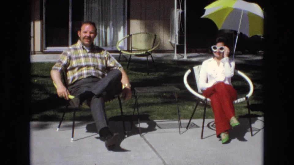 The man faces the camera while the woman sits with white sunglasses and red pants on a sunny residential patio in the 1973 California home-movie scene.