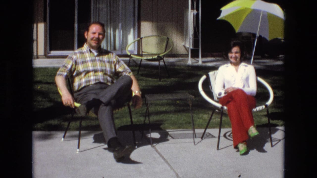 In a 1973 Central Coast California home-movie frame, a man in a plaid shirt and a woman in red pants sit in modern patio chairs on a sunny concrete patio.