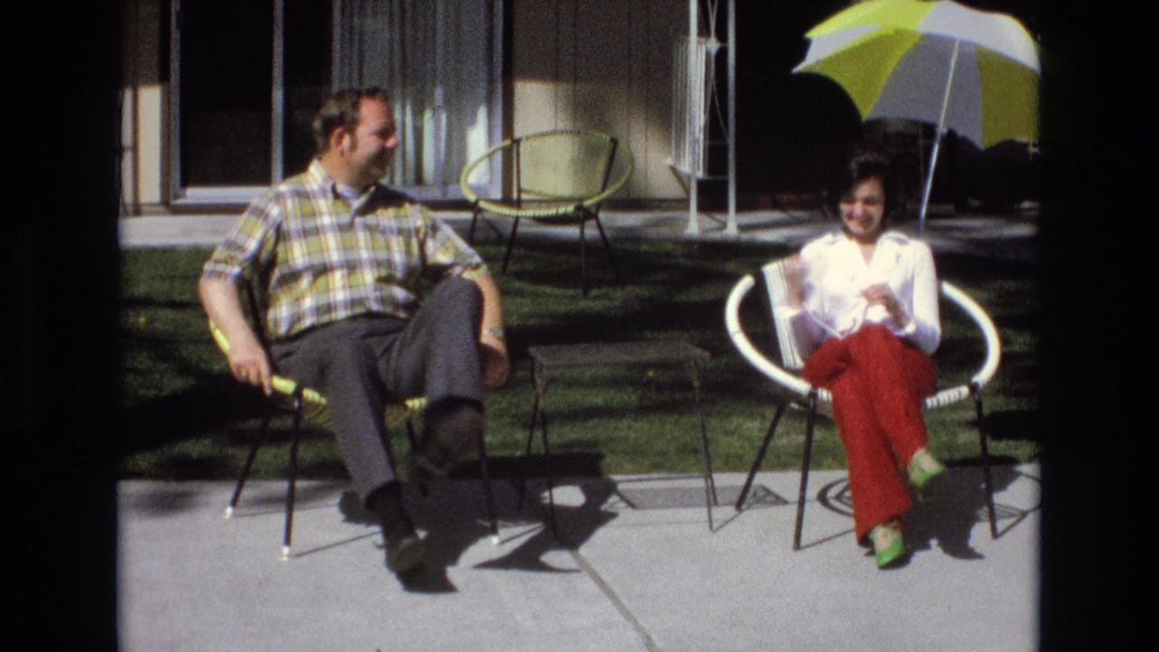 Seated man and woman smile on a patio beside green chairs and a striped umbrella.