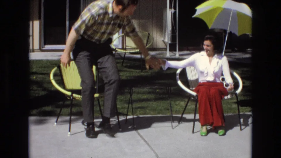 The man leans forward from a green patio chair and takes the seated woman by the hand as she prepares to stand, with the umbrella and house exterior still visible behind them.