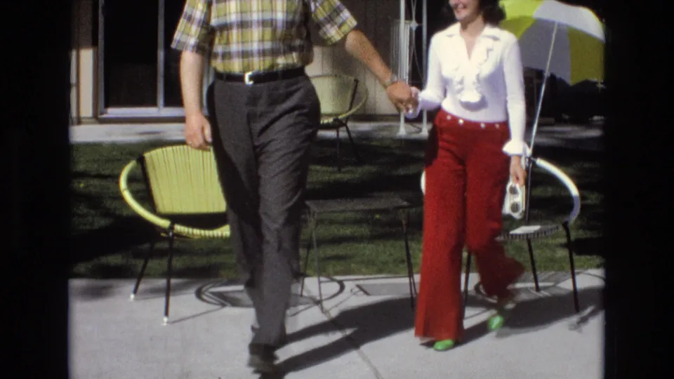 The couple walks hand in hand across the bright concrete patio, with the woman carrying a small object and the chairs, lawn, umbrella, and sliding door receding behind them.