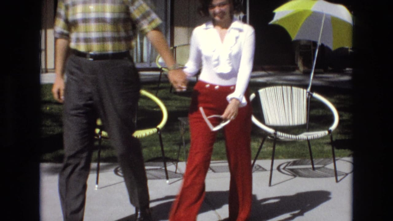 In a 1973 Central Coast California home-movie frame, an adult couple walks hand-in-hand across a sunlit patio with woven chairs and a yellow-and-white striped umbrella behind them.