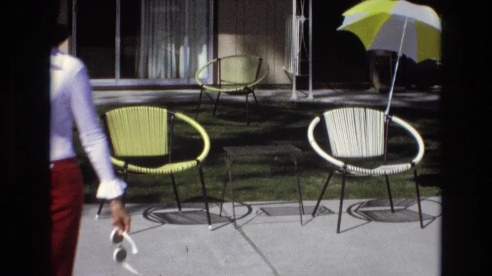 The woman in a white blouse and red flared pants moves past yellow and white woven patio chairs near a lawn and striped umbrella in the 1973 California home-movie scene.