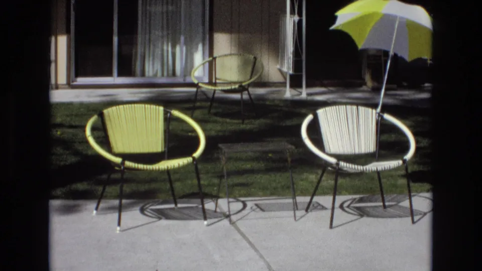 After the couple has passed, the frame holds on the sunlit patio furniture, including yellow and white woven chairs, a small table, lawn, sliding doors, and striped umbrella.