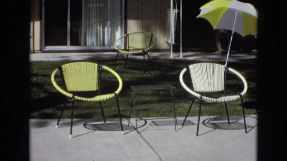 The final 1973 Central Coast California frame shows the patio after the couple has left the shot, with woven chairs, a small table, lawn, house exterior, and striped umbrella in sunlight.