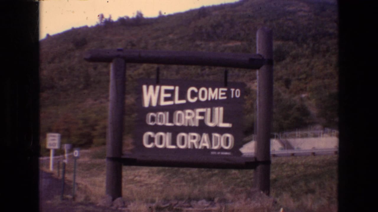 1973 Colorado 8mm frame showing a wooden Welcome to Colorful Colorado sign beside a roadside area with grassy ground and a tree-covered hill behind it.