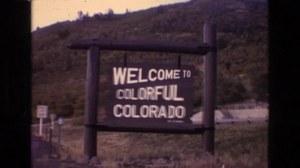 1973 Colorado 8mm frame showing a wooden Welcome to Colorful Colorado sign beside a roadside area with grassy ground and a tree-covered hill behind it.