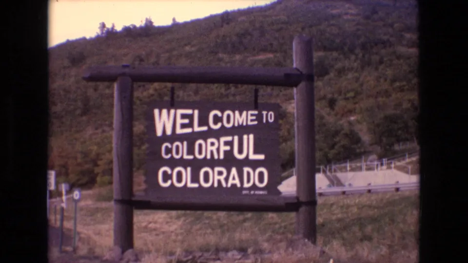 1973 Colorado 8mm frame with the state welcome sign centered against a green mountain slope and a pale roadside structure or guardrail in the background.