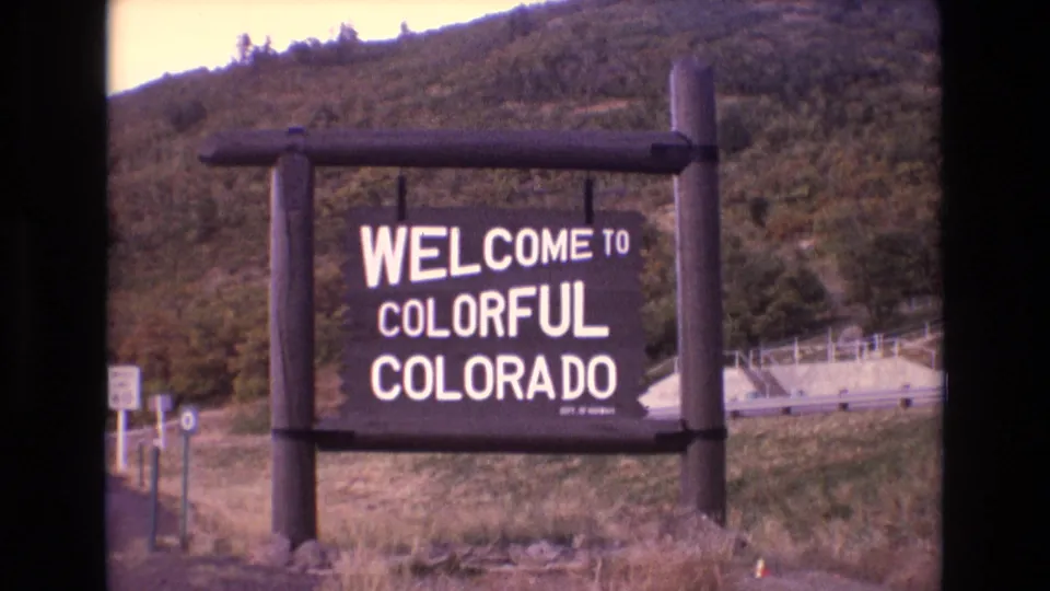 1973 Colorado home-movie frame showing the wooden state welcome sign from a visitor viewpoint, with grass, road edge, and wooded hillside visible around it.
