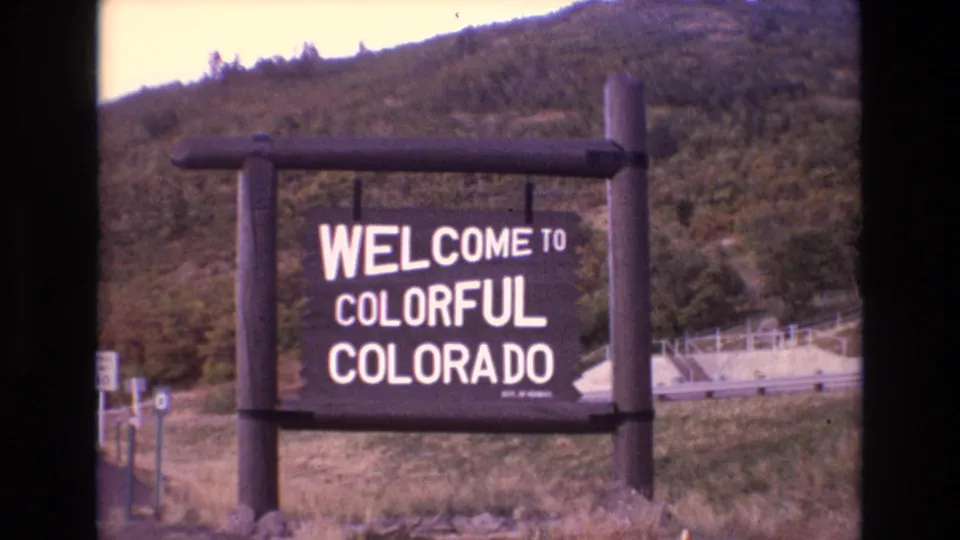 1973 Colorado 8mm frame holding on the Welcome to Colorful Colorado sign, with wooden supports, roadside vegetation, and a mountain hillside in the background.