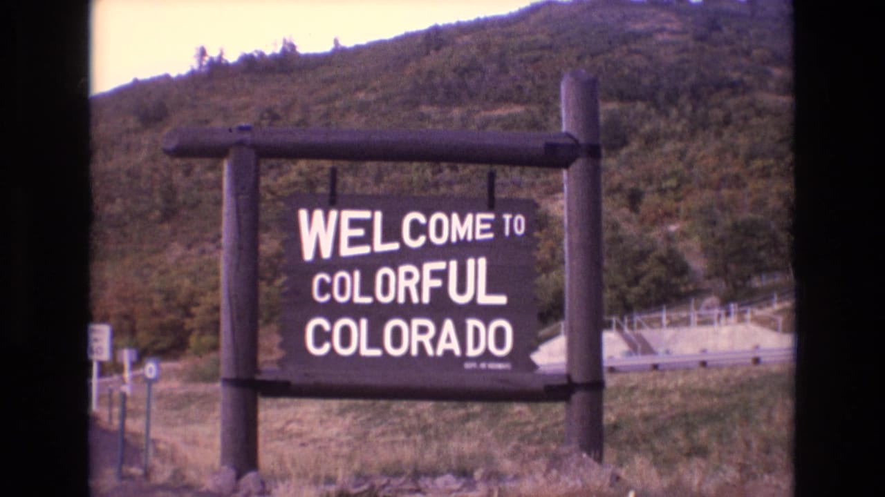 1973 8mm Colorado travel frame showing a wooden Welcome to Colorful Colorado roadside sign against a forested hillside, with dark film borders visible.