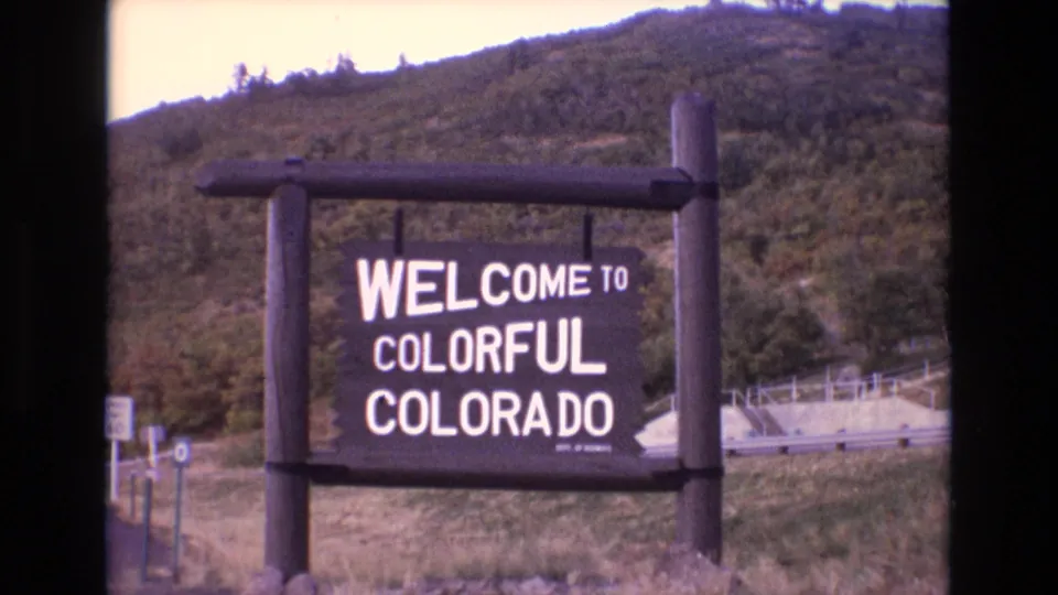 1973 Colorado home-movie frame showing the state welcome sign in front of a green hillside, with 8mm softness and dark border areas.