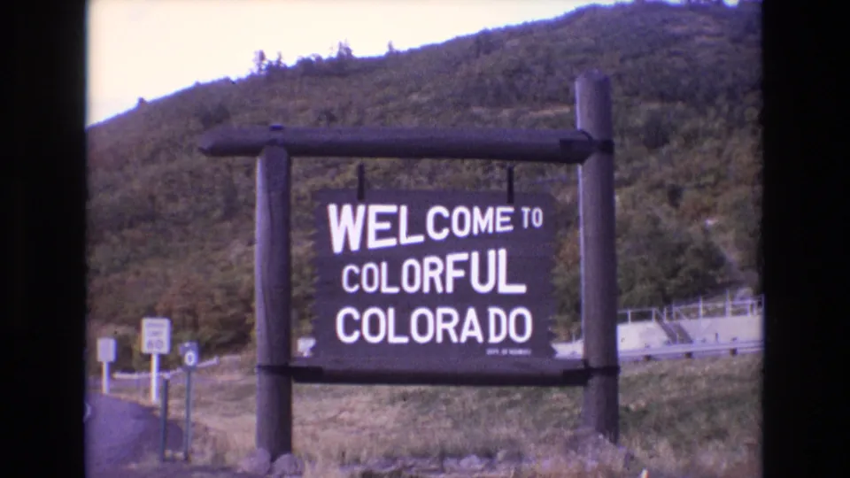 Home-movie frame of the Welcome to Colorful Colorado sign on a rustic wooden support, with grass, guardrail, and a wooded hillside behind it.